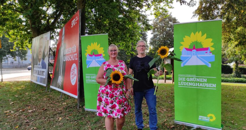 Foto v.L.n.R.: Anke Brandmeier, Lars Reichmann - auf dem Bild sieht man, wie die beiden am Kreisverkehr vor Roll-Ups und mit Blumen in der Hand stehen.